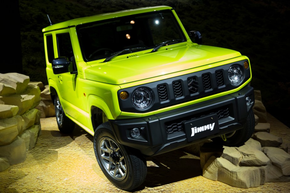 A lime green Suzuki Jimny parked on a rocky off-road obstacle on stage at a foreign market auto show.