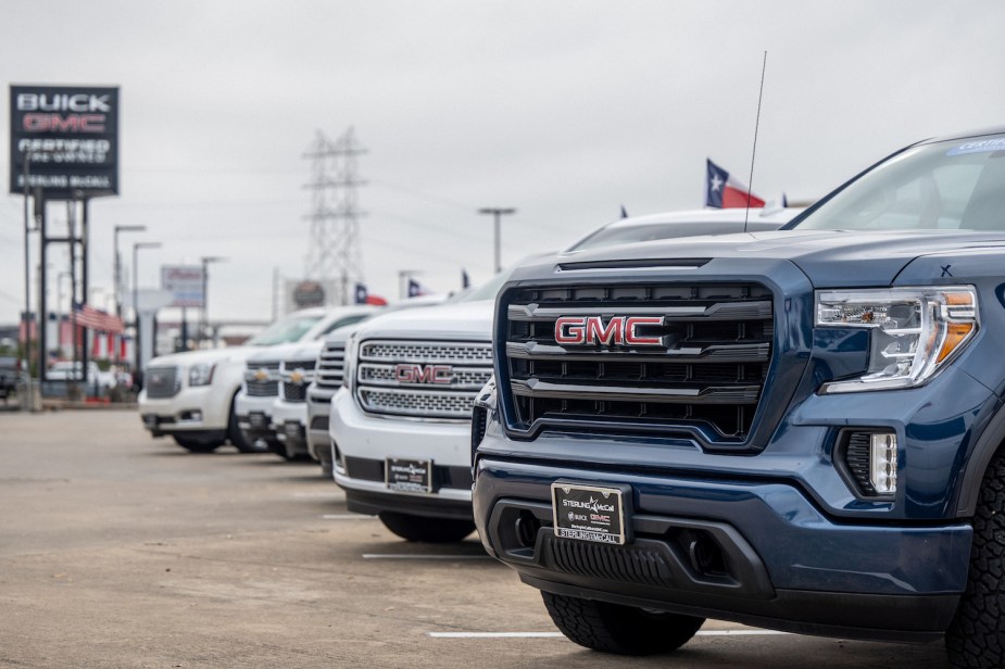 A row of both GMC and Chevrolet pickup trucks parked at a General Motors dealership, a Texas flag visible in the background.