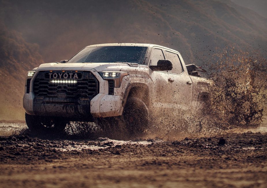 A white 4WD Toyota Tundra pickup truck drives through mud, kicking up dirt with mountains visible in the background.