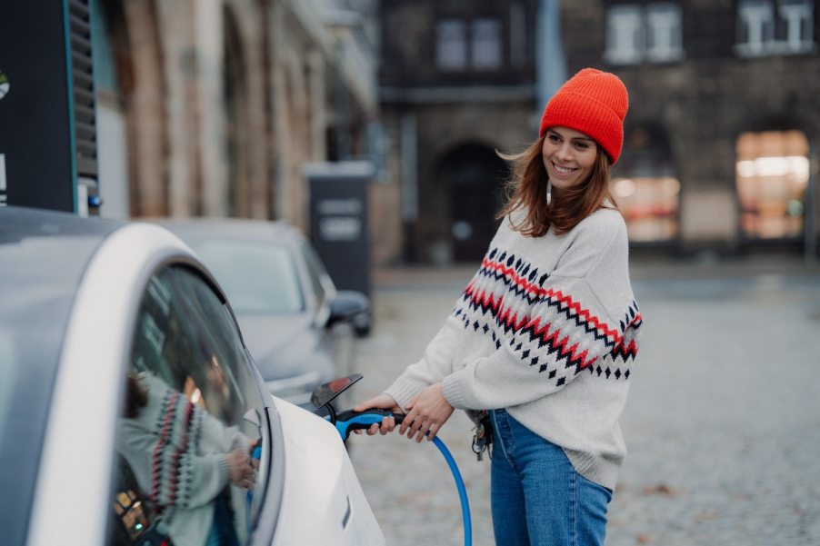 A woman in an orange hat plugs a rapid charger cable in to the port of her Tesla sedan.