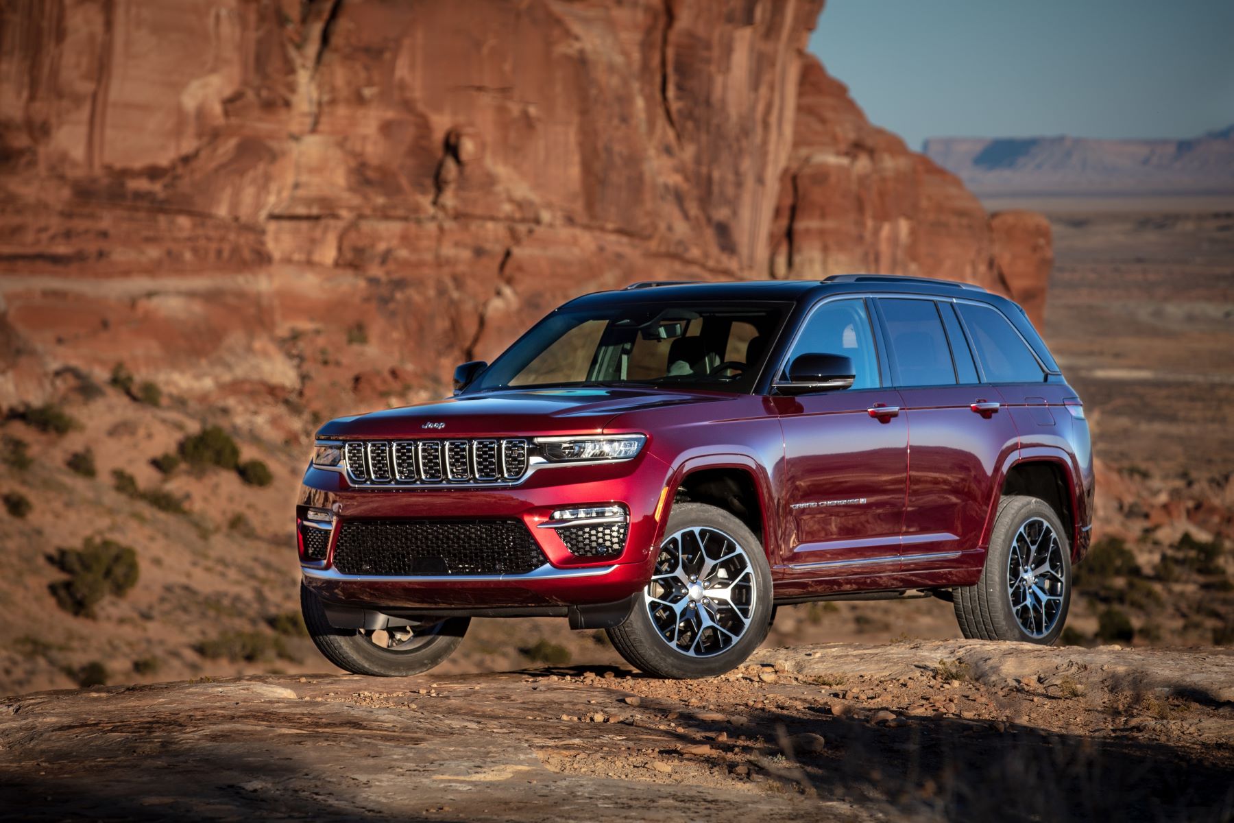 A red 2023 Jeep Grand Cherokee Summit Reserve midsize SUV model parked on rocky terrain in a desert near red cliffs