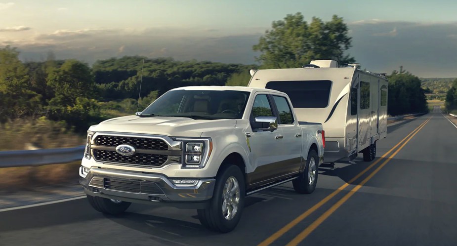 A white 2023 Ford F-150 full-size pickup truck is driving on the road towing a trailer.