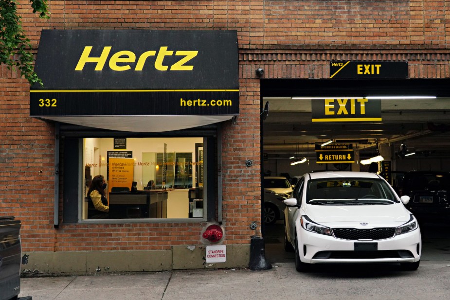 White Kia rental car driving out of a Hertz branch.