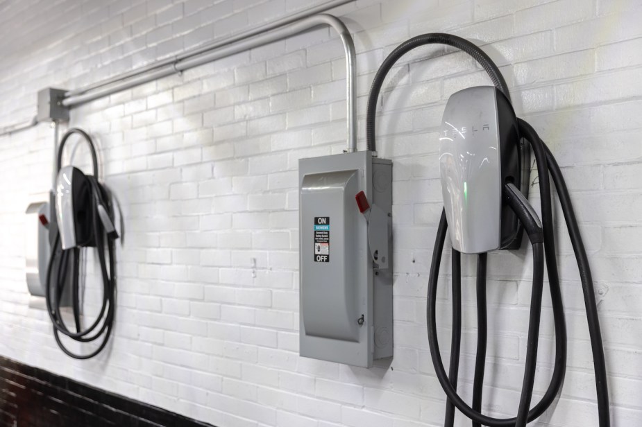Two home EV chargers hung from a white brick wall and wired to a breaker panel.