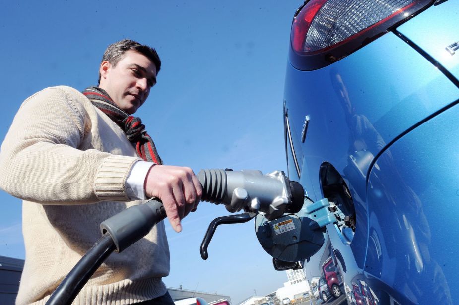 A man plugs a charging cable into the port of his blue electric vehicle, the sky visible behind him.