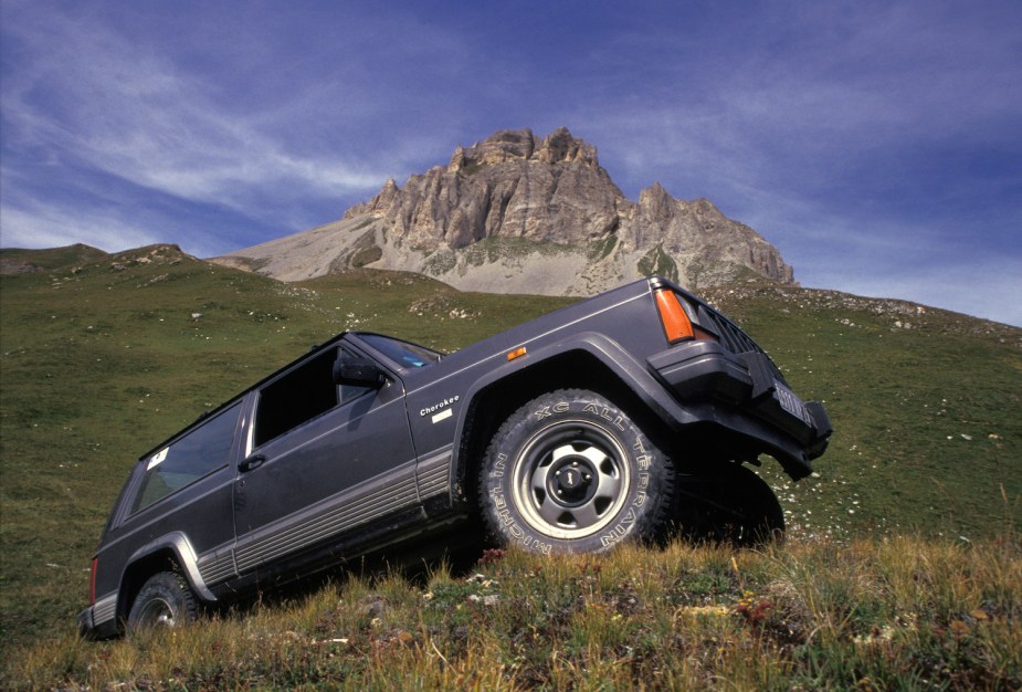 A gray Jeep Wrangler 4x4 SUV parked on a grassy hillside, rocky outcroppings visible in the background.