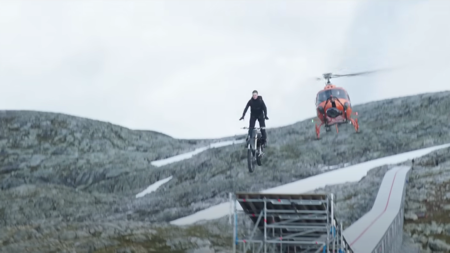 Actor Tom Cruise rides a motorcycle off a clifftop ramp for a Mission Impossible stunt, a helicopter visible in the background.