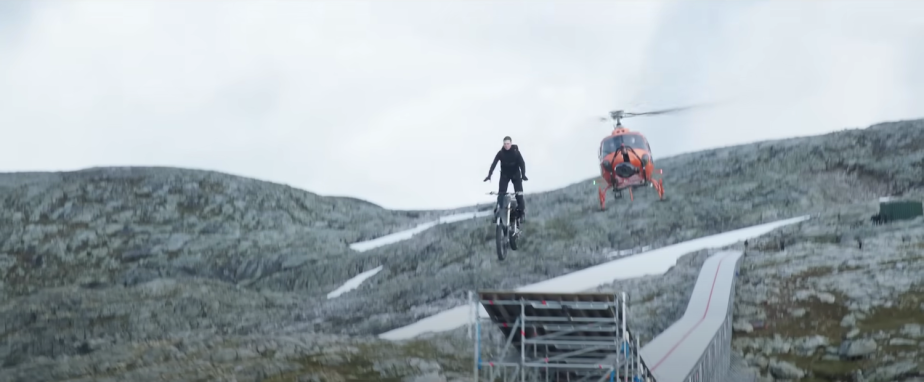 Actor Tom Cruise rides a motorcycle off a clifftop ramp for a Mission Impossible stunt, a helicopter visible in the background.