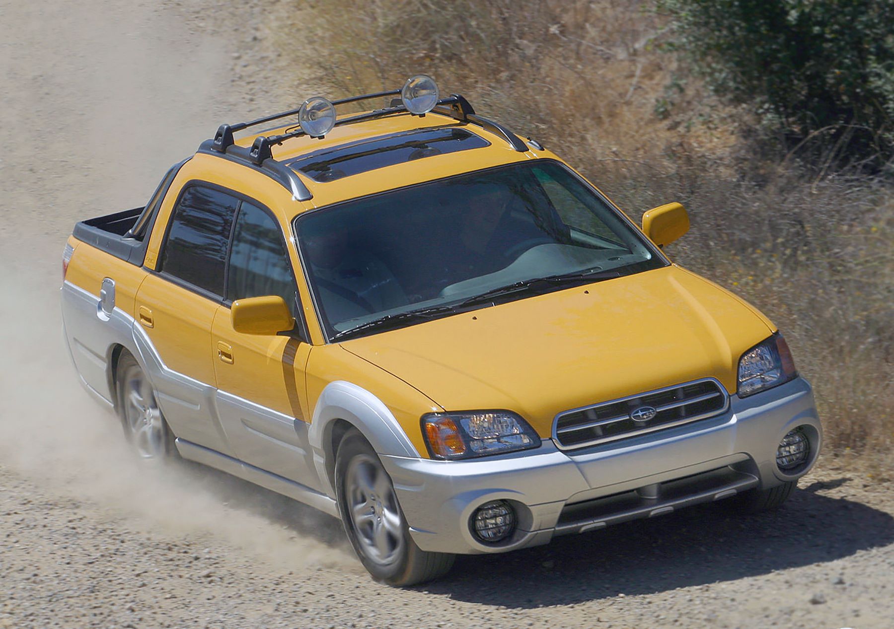 A yellow Subaru Baja compact utility pickup truck built with a midgate driving on a dusty gravel road