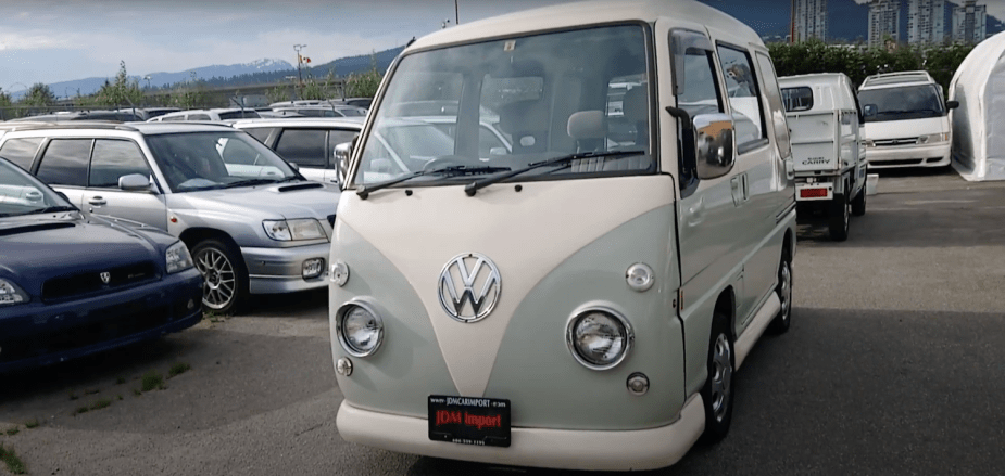 A green and cream VW bus lookalike Subaru Sambar parked on a JDM import car lot, Kei trucks in the background.