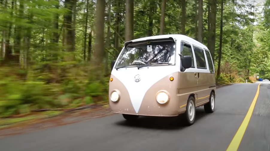 A brown Subaru Sambar converted into a Volkswagen bus lookalike drives on a rural road through pine tree forests.