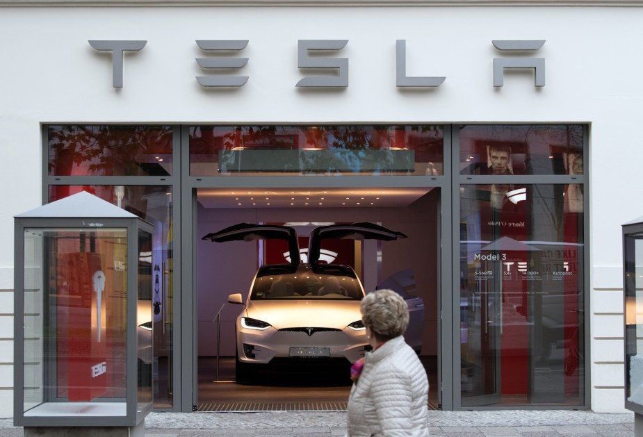 A person stands outside a Tesla showroom, looking at an electric SUV parked inside.