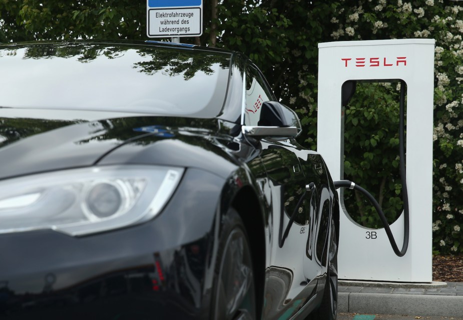 Closeup of a black Tesla vehicle plugged into a super charger, trees visible in the background.