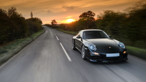 A modified Porsche 997 Carrera S sports car driving on a rural road with a sunset visible in the background.