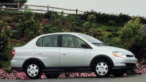 A light-gray 2000-2002 first-generation Toyota Echo sedan parked by pink flowers