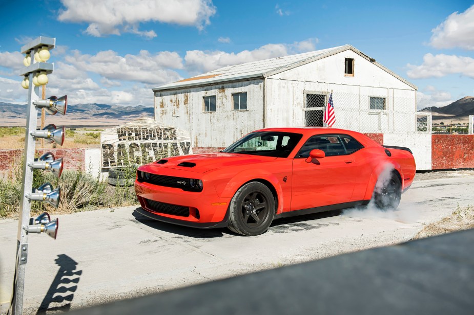 An orange Dodge Challenger SRT Super Stock with a Hellcat motor burns tire in front of a raceway sign.