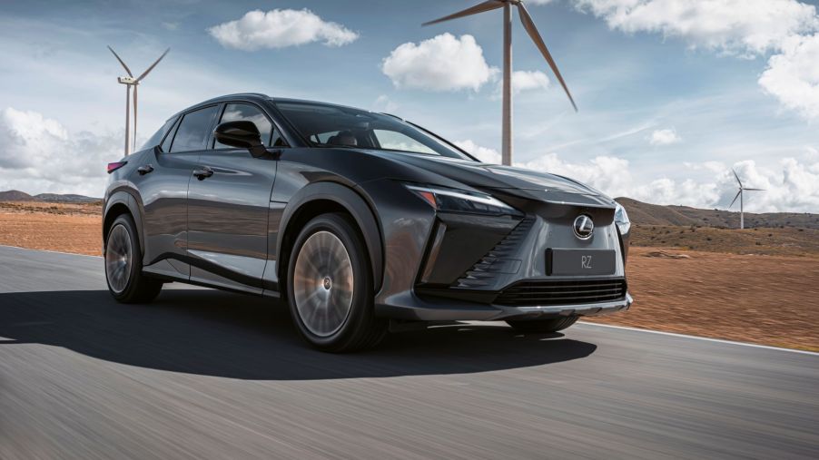 A dark-gray 2023 Lexus RZ all-electric luxury SUV driving on a highway past windmills