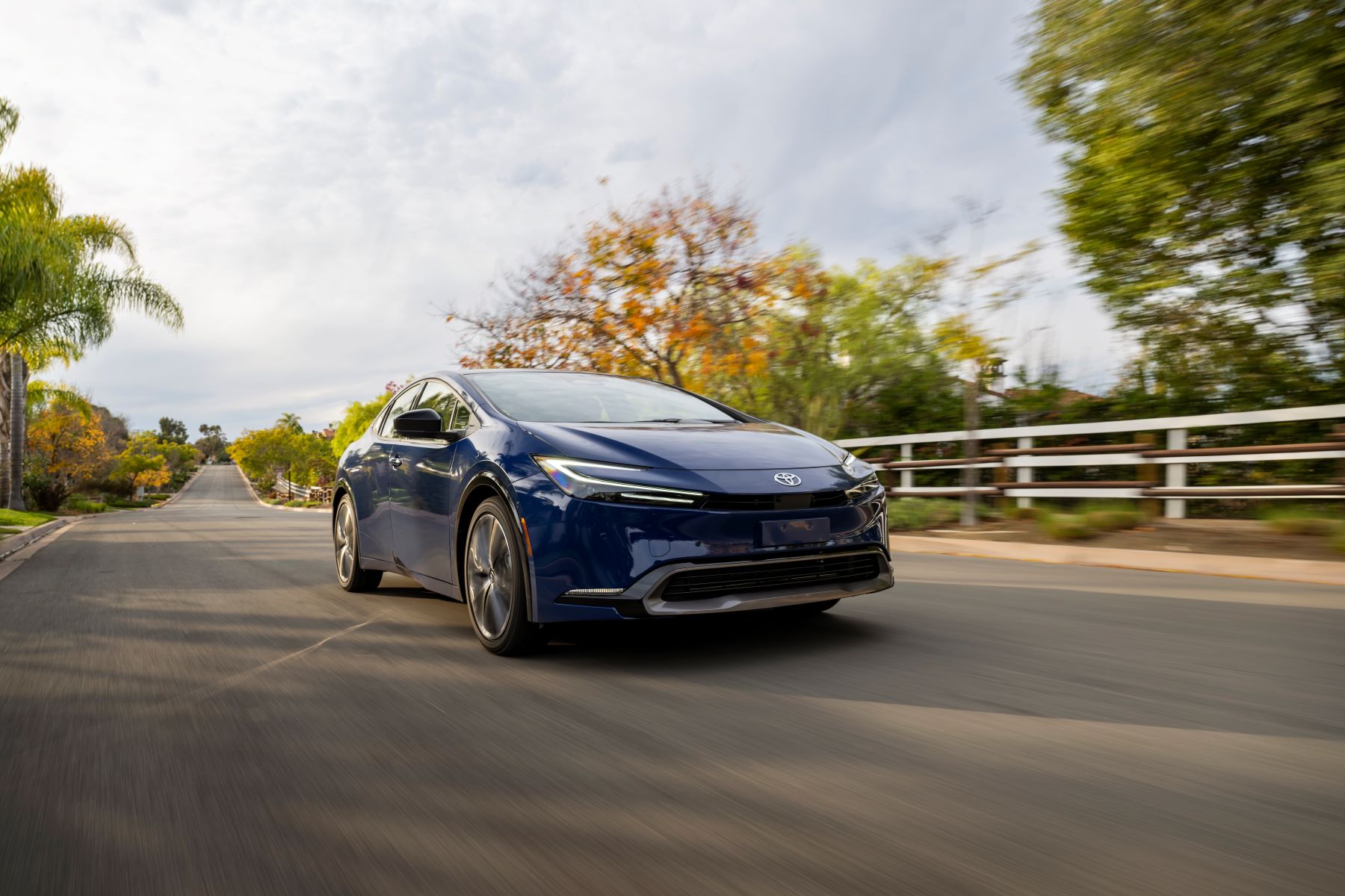 A 2023 Toyota Prius Limited in Reservoir Blue driving past white fencing on a country asphalt road