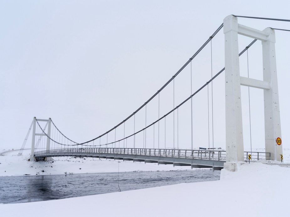 A suspension bridge spanning the snow covered banks of a icy river in winter.