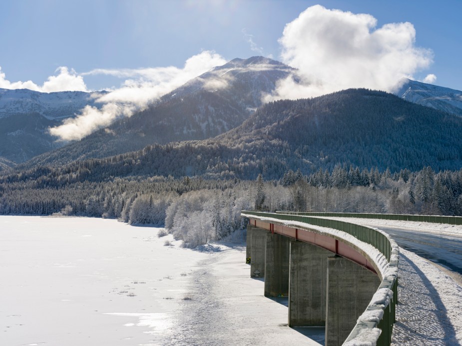 A bridge winds across a frozen pond and into cold, wintertime snowy mountains.