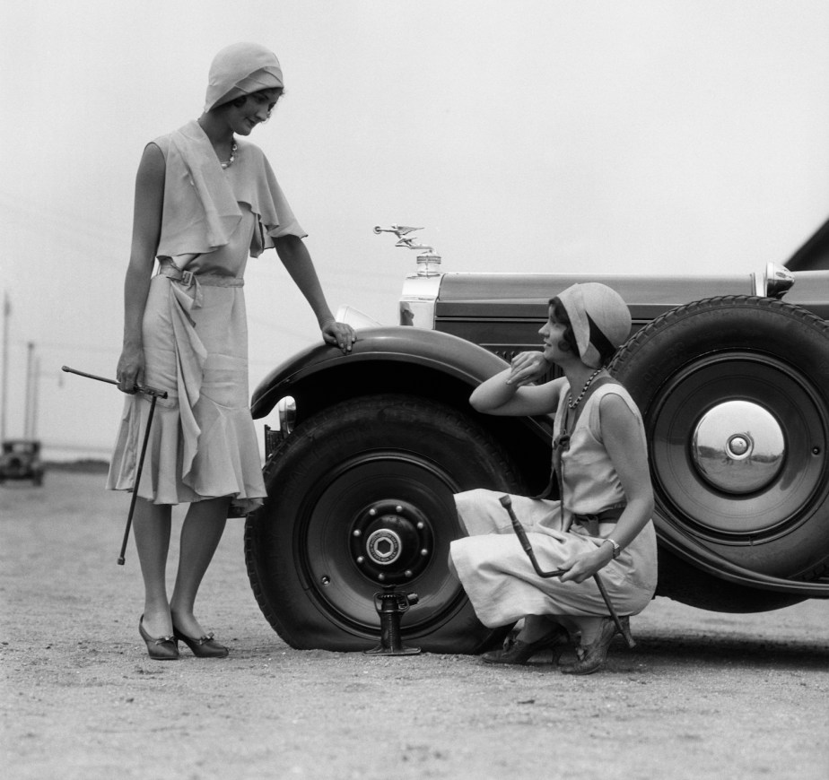 Two women in dresses pose by a car with a flat tire, halfway through a tire change in 1937.