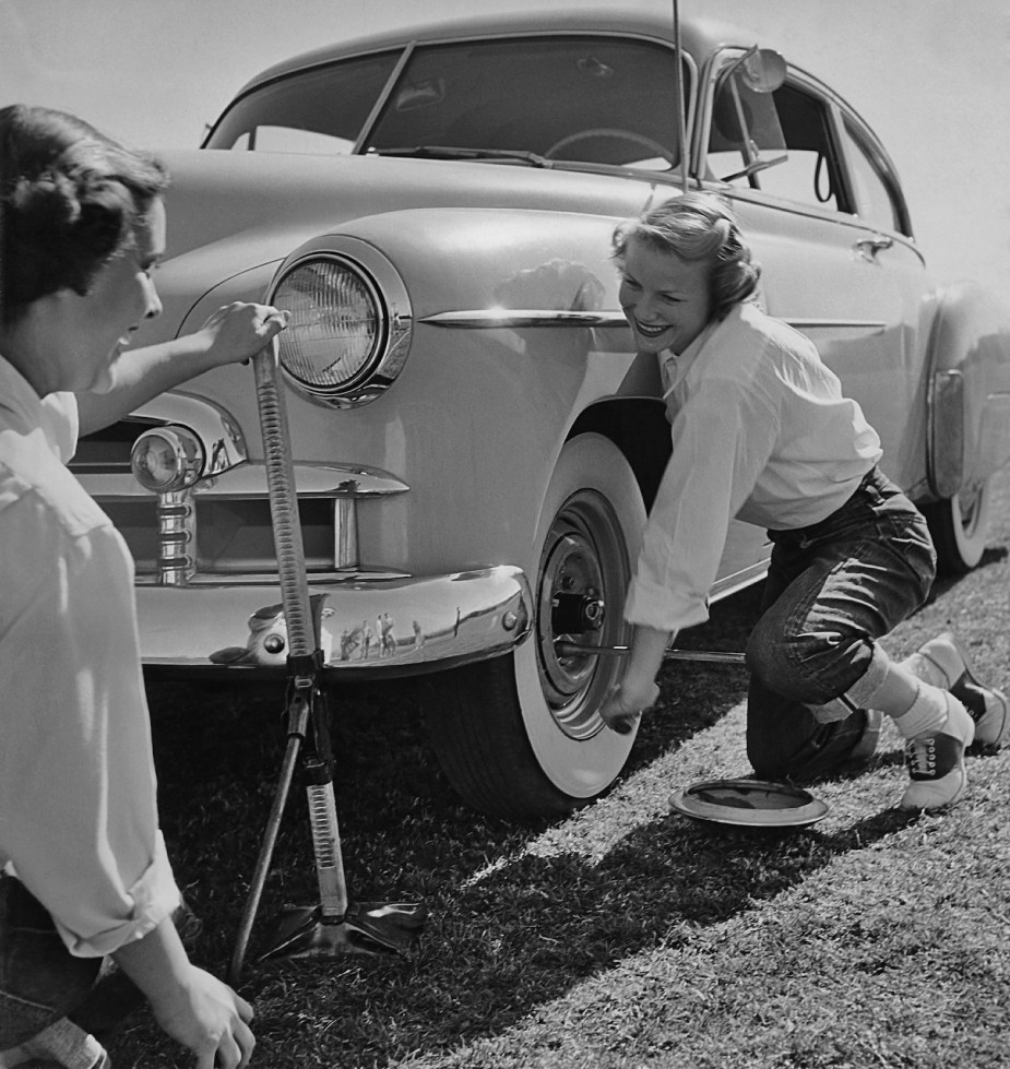 Black and white photo of two women competing in a tire change speed contest in the 1950s, one of them operating a jack and the other spinning the lug wrench.