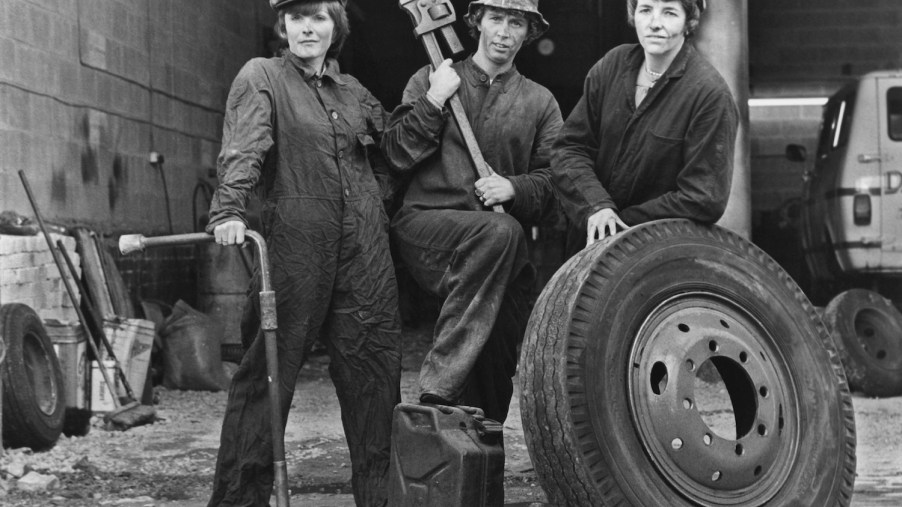 Three 1960s female mechanics pose for a black and white photo with a truck tire, a pipe wrench, and a lug wrench with a breaker bar extension to break nuts free.