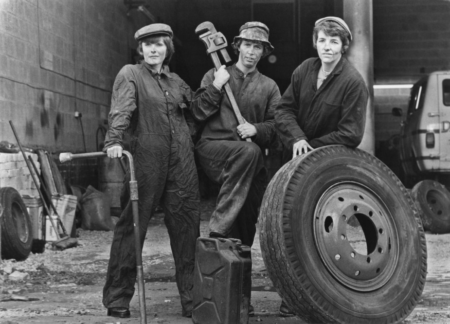 Three 1960s female mechanics pose for a black and white photo with a truck tire, a pipe wrench, and a lug wrench with a breaker bar extension to break nuts free.