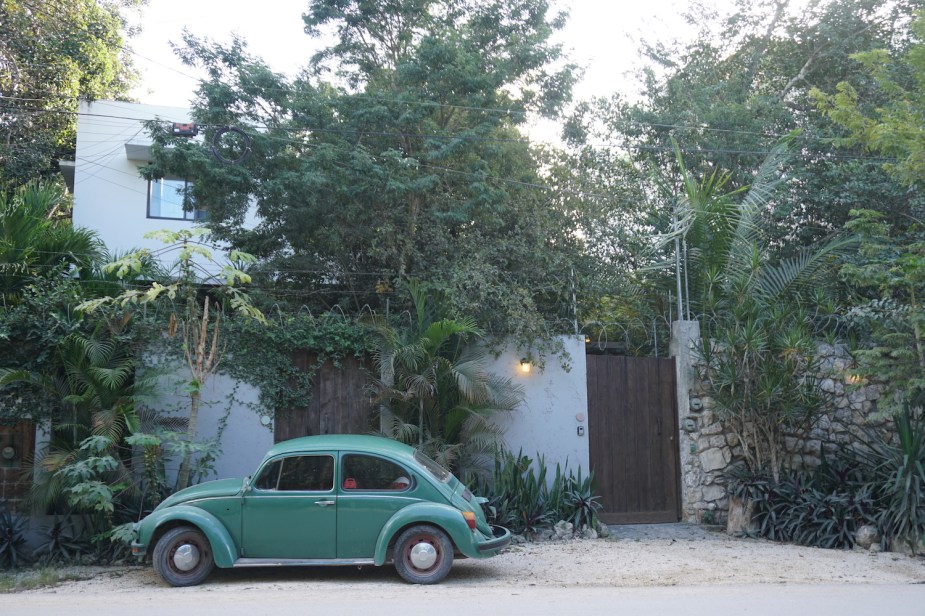 A green VW Beetle parked on a street in Mexico, a house and tropical treeline visible in the background.