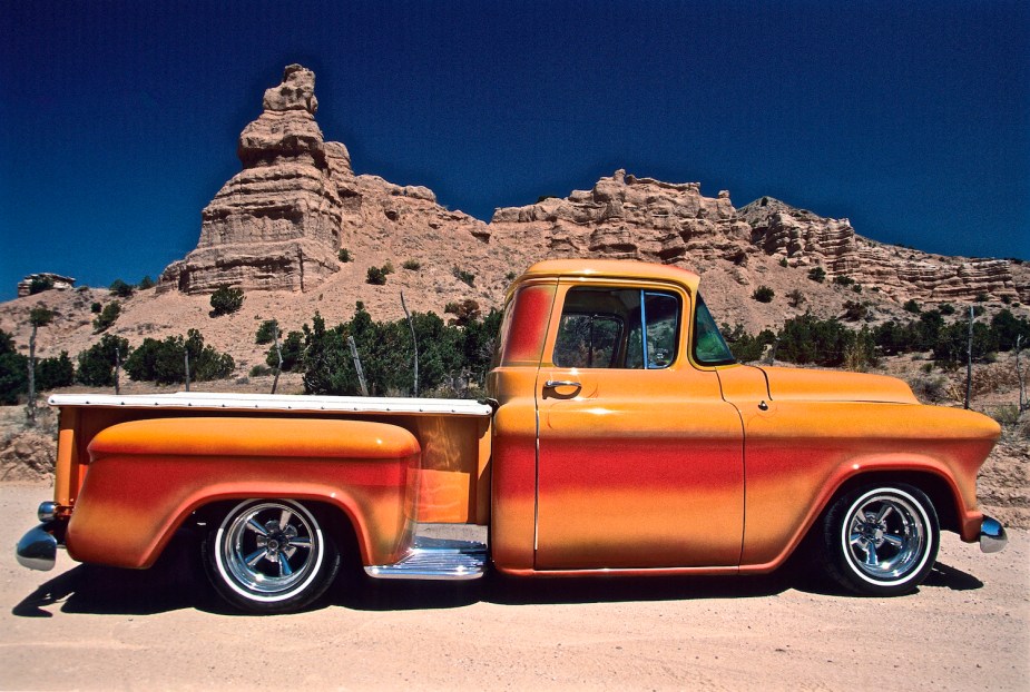 This orange pickup truck is dropped considerably, and parked in a desert in front of some cliffs.
