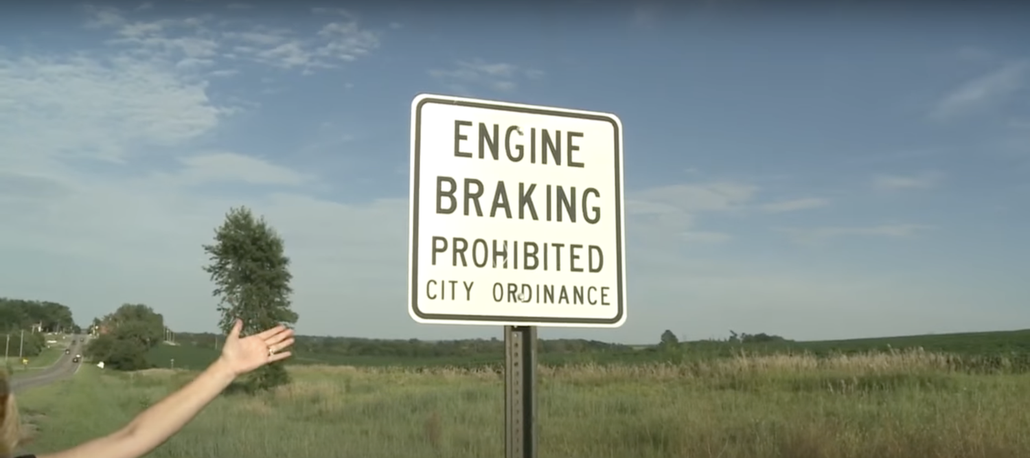 A white "Engine Braking Prohibited: City Ordinance" road sign with a grassy field visible behind it.