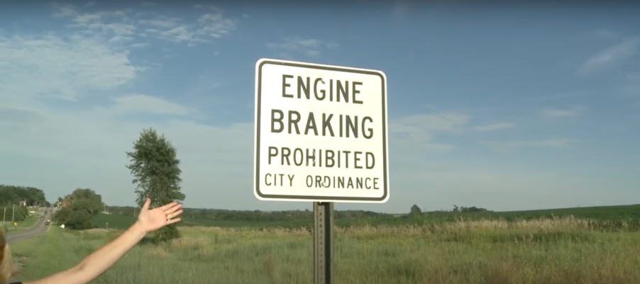 A white "Engine Braking Prohibited: City Ordinance" road sign with a grassy field visible behind it.