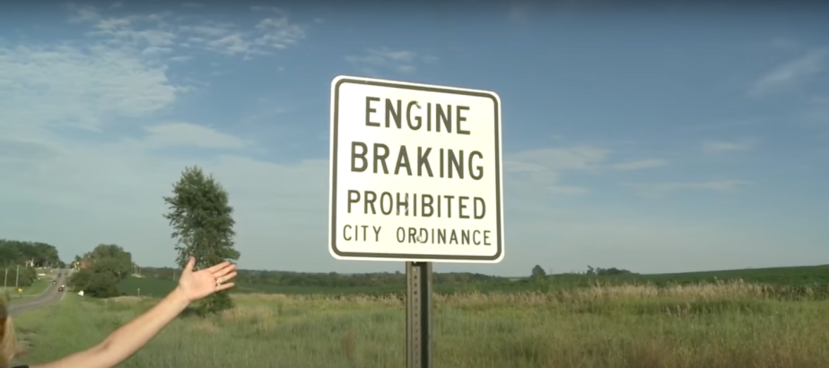 An "Engine Braking Prohibited" sign on a roadside, a grassy field visible in the background.