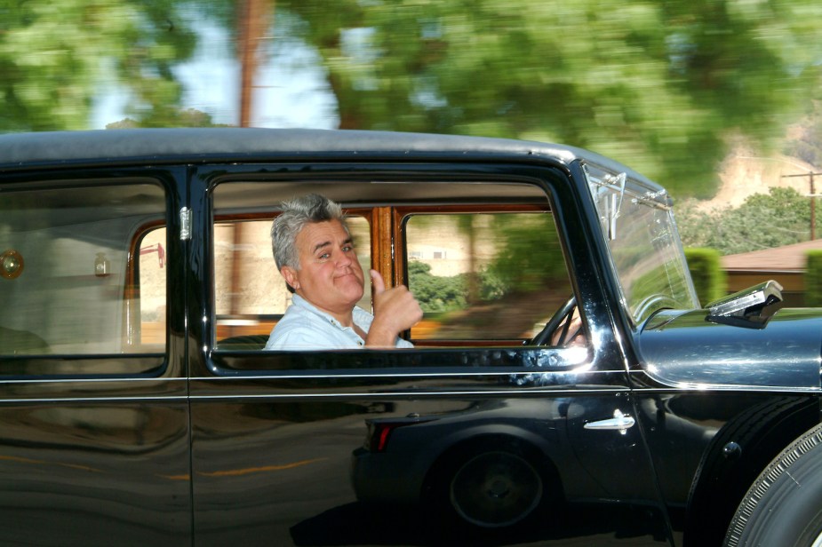 Jay Leno flashes a thumbs up while driving a classic collector's car.