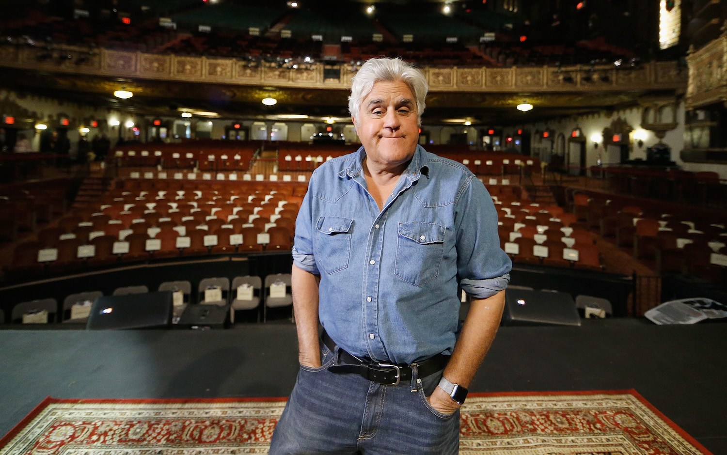 Jay Leno stands on a stage before a comedy show, the audience chairs visible behind him.