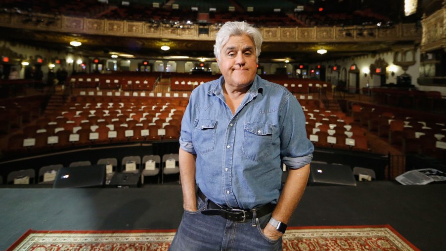 Jay Leno stands on a stage before a comedy show, the audience chairs visible behind him.