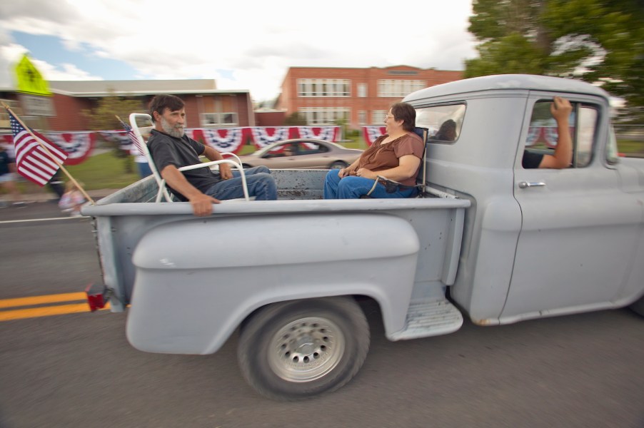 A couple rides in the bed of a primer gray stepside pickup truck during a parade.
