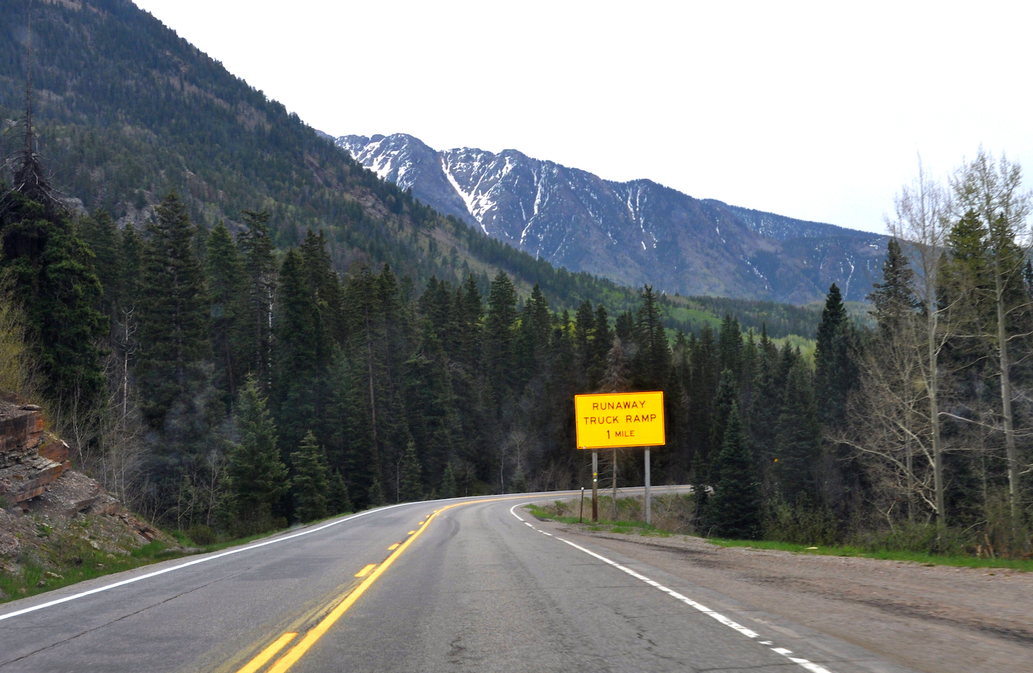 A bright yellow "Runaway Truck Ramp 1 Mile" sign by the side of a road in front of the mountains of Colorado.