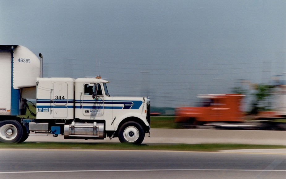 A white runaway semi truck tractor speeding down the road, an orange truck and telephone poles blurring by in the background.