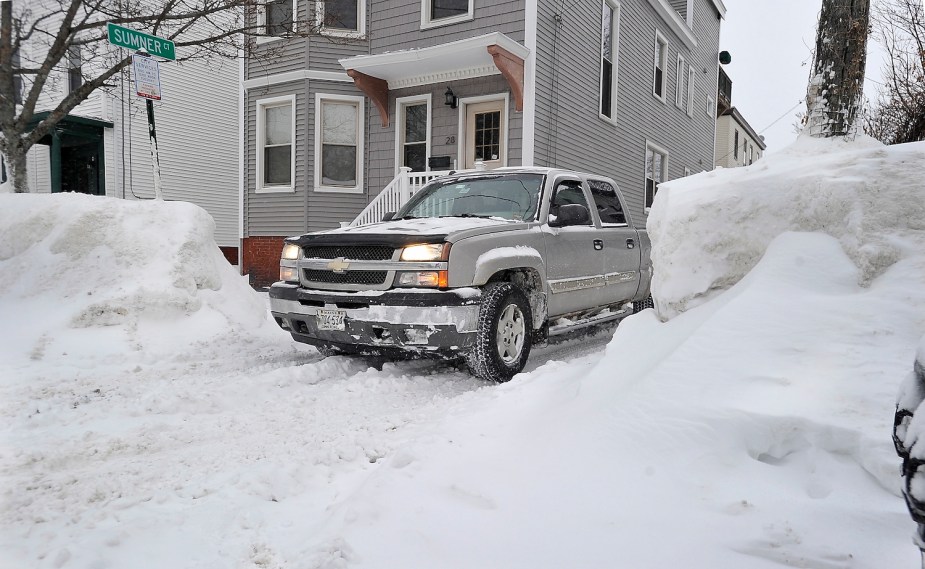 A gray truck driving down a driveway, between tall snowbanks, a house visible in the background.