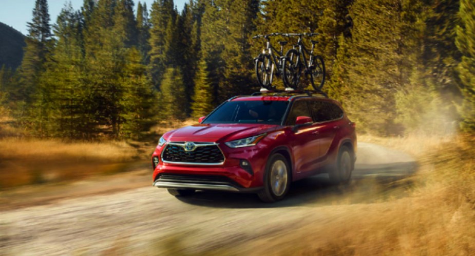 A red 2023 Toyota Highlander Hybrid midsize hybrid SUV is driving on the road. 