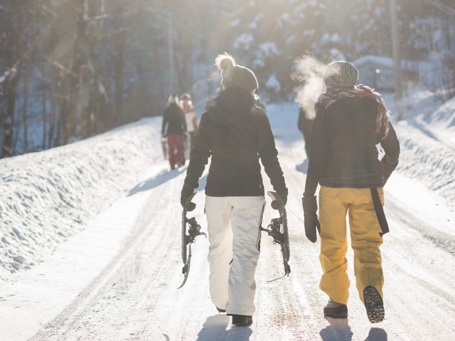 Women in snow gear walking down a winter road, carrying snowshoes.