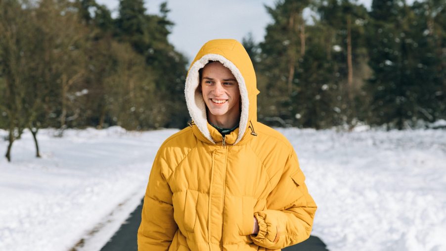 A person walking down a snowy road in a warm yellow parka jacket, trees visible in the background.