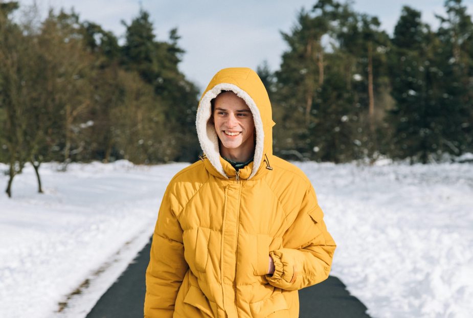 A person walking down a snowy road in a warm yellow parka jacket, trees visible in the background.