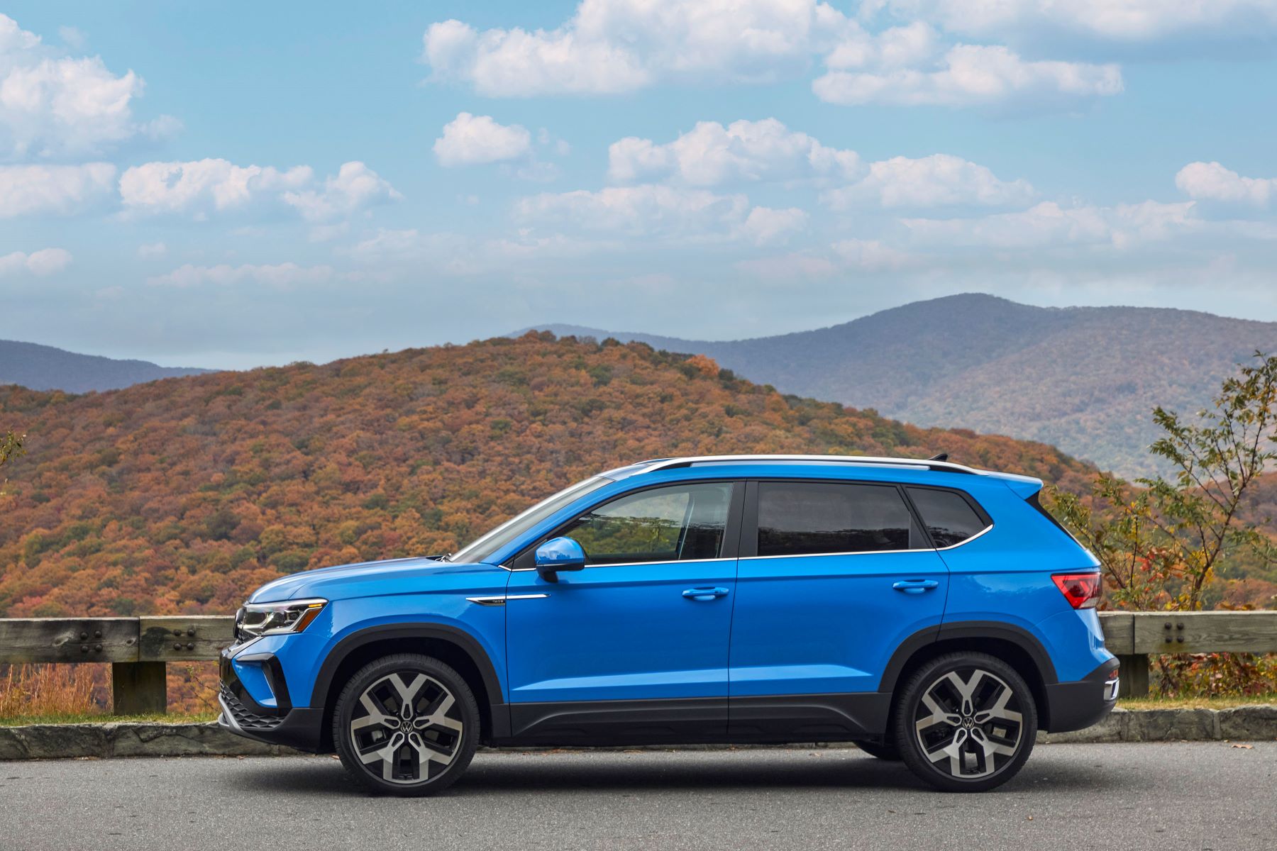 A side profile shot of a blue 2022 Volkswagen Taos compact SUV model parked near wooden fencing near forest hills