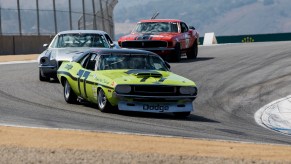 A limelight green 1970 Dodge Challenger leads a Camaro and Mustang during a trans america reenactment race on a track, mountains visible in the background.