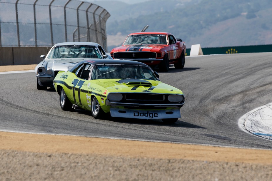 A limelight green 1970 Dodge Challenger leads a Camaro and Mustang during a trans america reenactment race on a track, mountains visible in the background.