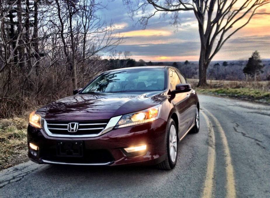 A maroon 2013 Honda Accord midsize car shows off its lights while it drives down a country road. 