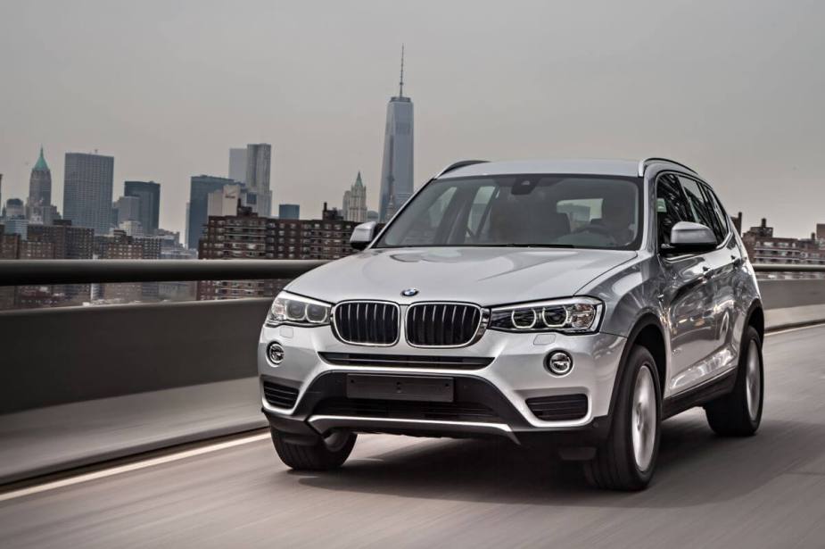 A silver-gray 2014 BMW X3 xDrive luxury compact SUV model driving on a highway with an urban city in the background