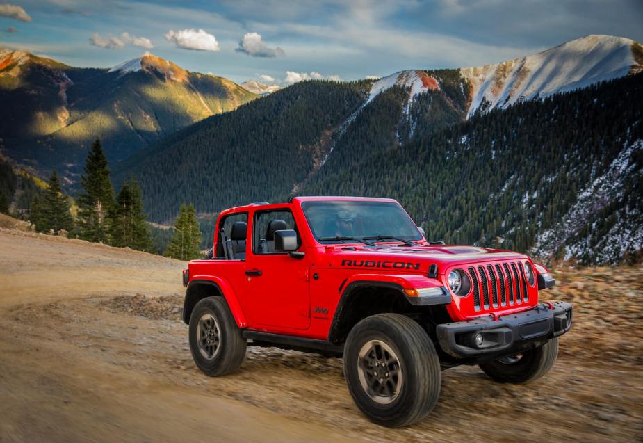 Bright red Jeep Wrangler Rubicon convertible SUV with its top down navigates a switchback in a dirt trail, snow-capped mountains visible in the background.